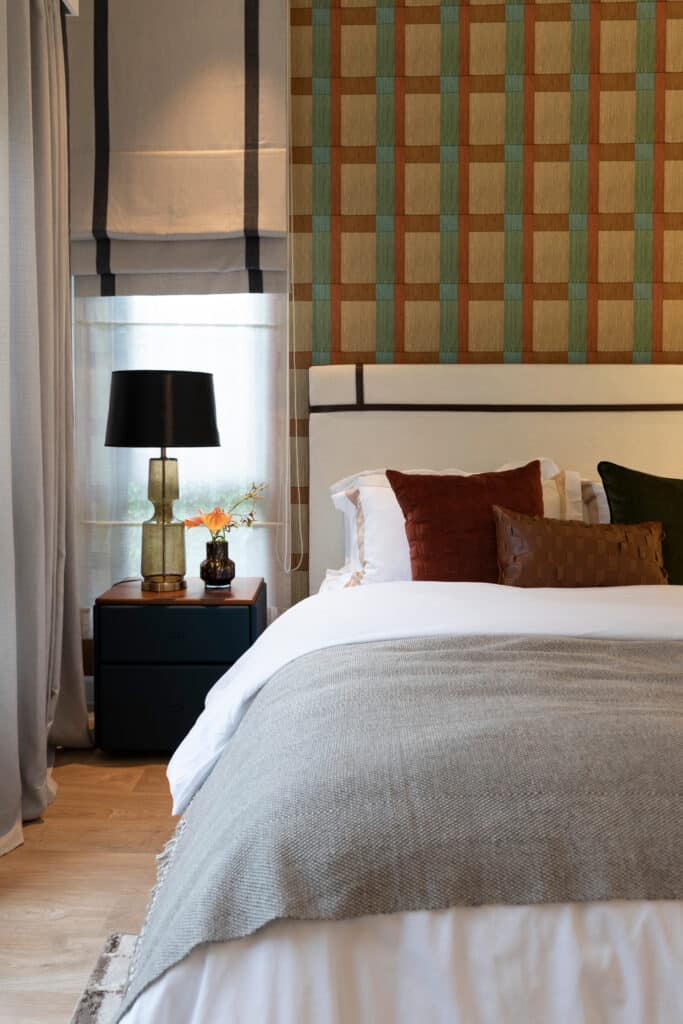 Chambre moderne avec lit blanc, jeté gris, coussins roux et verts. Papier peint géométrique ocre, vert, orange. Chevet foncé, lampe design.