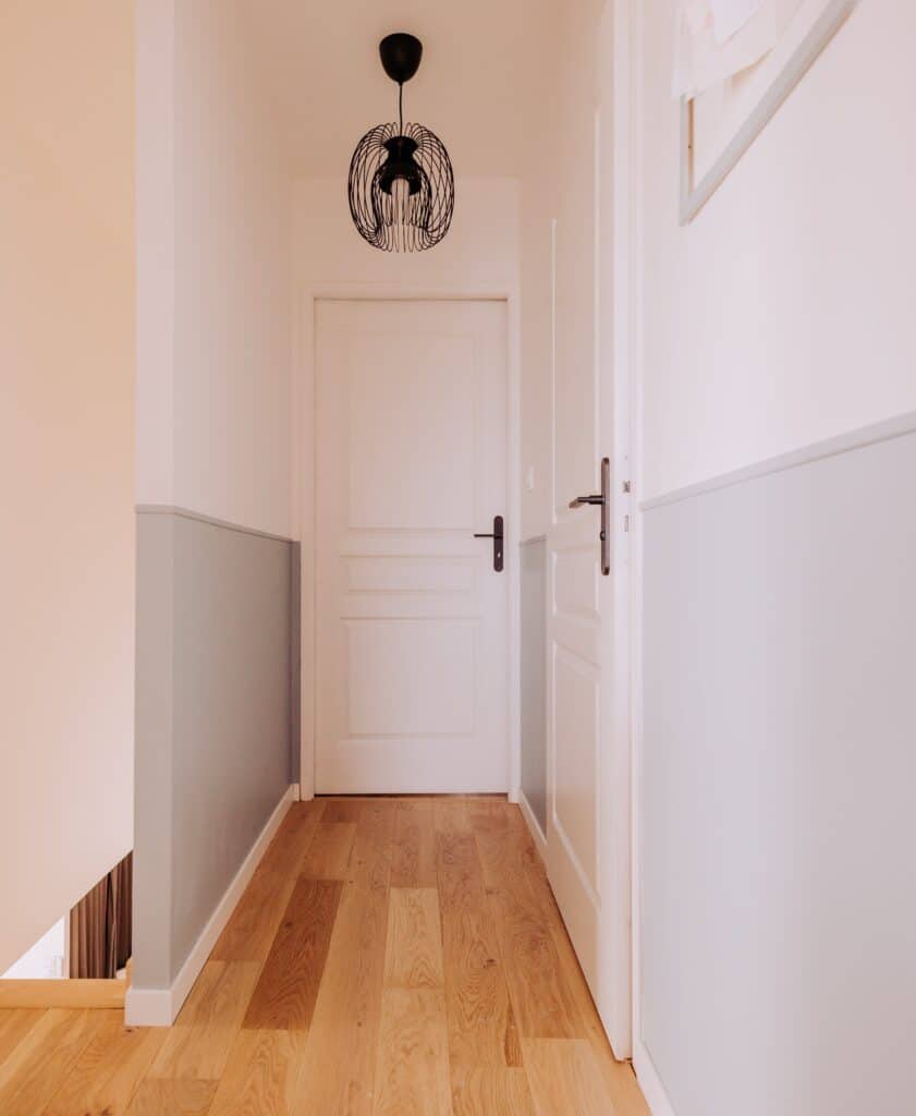 Couloir moderne avec parquet en bois clair, murs blancs et lambris gris. Deux portes blanches et un luminaire suspendu noir.