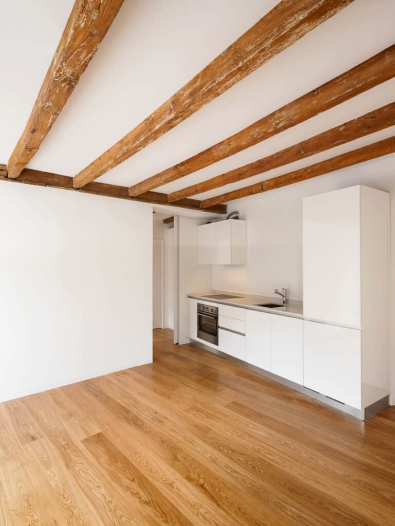 Cuisine moderne blanche épurée avec four et évier inox, parquet en bois clair et plafond avec poutres apparentes dans une pièce lumineuse.