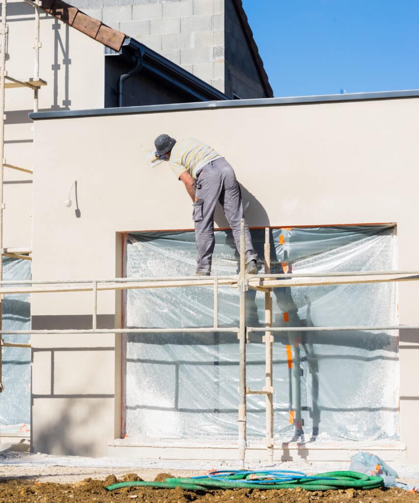 Ouvrier du bâtiment appliquant de l'enduit sur la façade d'une maison neuve en construction, avec échafaudage et fenêtres bâchées sous un ciel bleu.