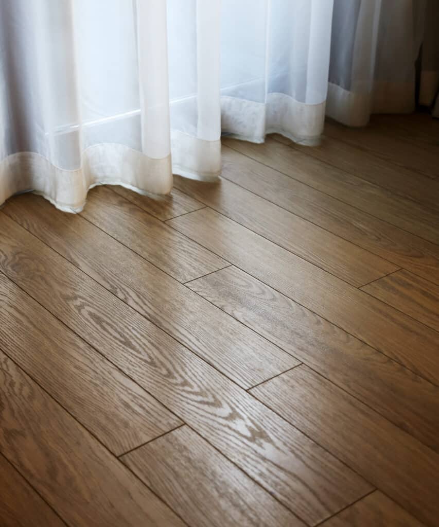Sol en parquet clair avec motif grain de bois, baigné de lumière naturelle filtrant à travers de longs rideaux blancs devant une fenêtre.