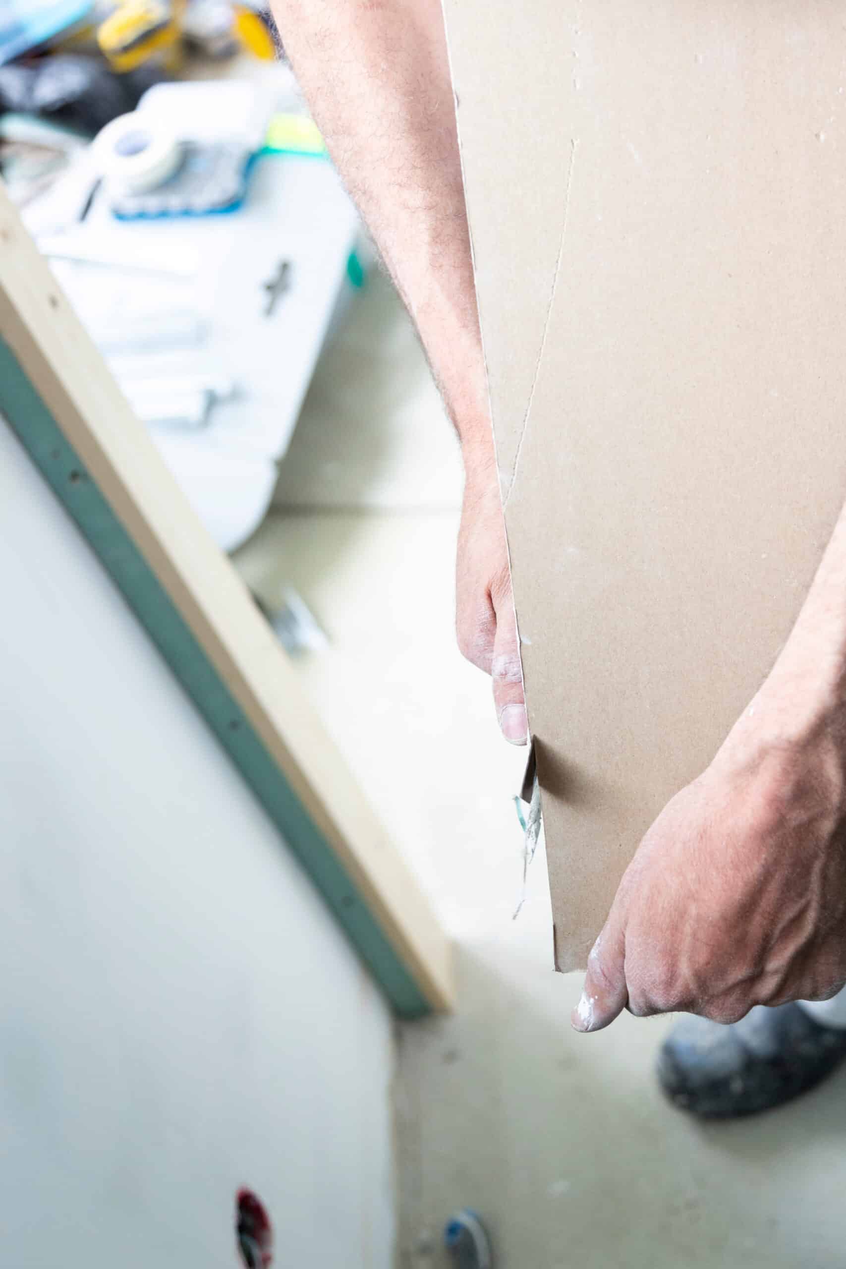 Mains poussiéreuses d'un plaquiste portant une planche de placoplâtre sur un chantier de rénovation.