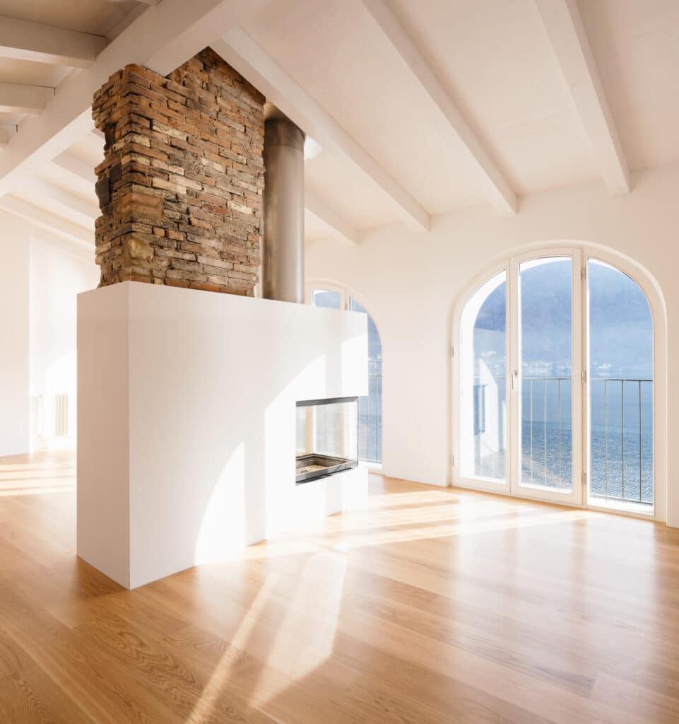 Grand salon lumineux avec cheminée pierre/moderne, plancher bois et poutres blanches. Fenêtres arquées sur lac et montagnes ensoleillées.