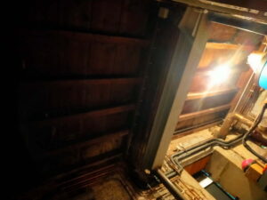 Gros plan sur un sous-sol ou une cave sombre avec poutres en bois apparentes, une colonne de support grise, et des canalisations de cuivre et câbles exposés éclairés par une lumière vive.