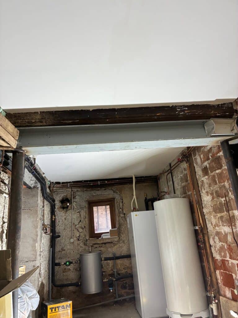 Local technique sous poutre métallique et bois, avec chaudière ou pompe à chaleur blanche et réservoirs, murs en pierre brute et tuyauterie apparente. Boîte TITAN visible.
