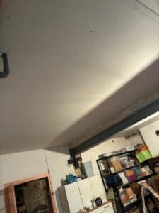 Plafonnier en plaques de plâtre neuves avec vis apparentes, conduit de tuyauterie visible, poutre métallique et espace de rangement désordonné en bas.