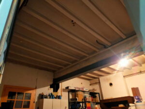 Plafond intérieur avec poutres apparentes, solives en bois claires et une poutre de soutien métallique foncée, éclairage puissant créant un reflet.