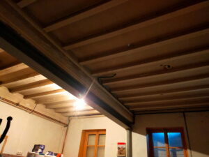 Plafond intérieur rustique avec poutre métallique noire et solives en bois apparentes, éclairage vif près de la poutre, fenêtres en bois.