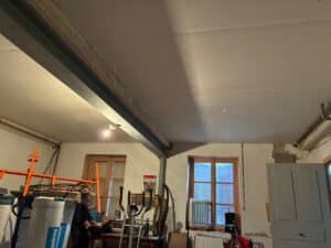 Intérieur en cours de rénovation, plafond en plaques de plâtre avec poutre métallique apparente, fenêtre à petits carreaux, outils et matériaux (isolation KNAUF) visibles.
